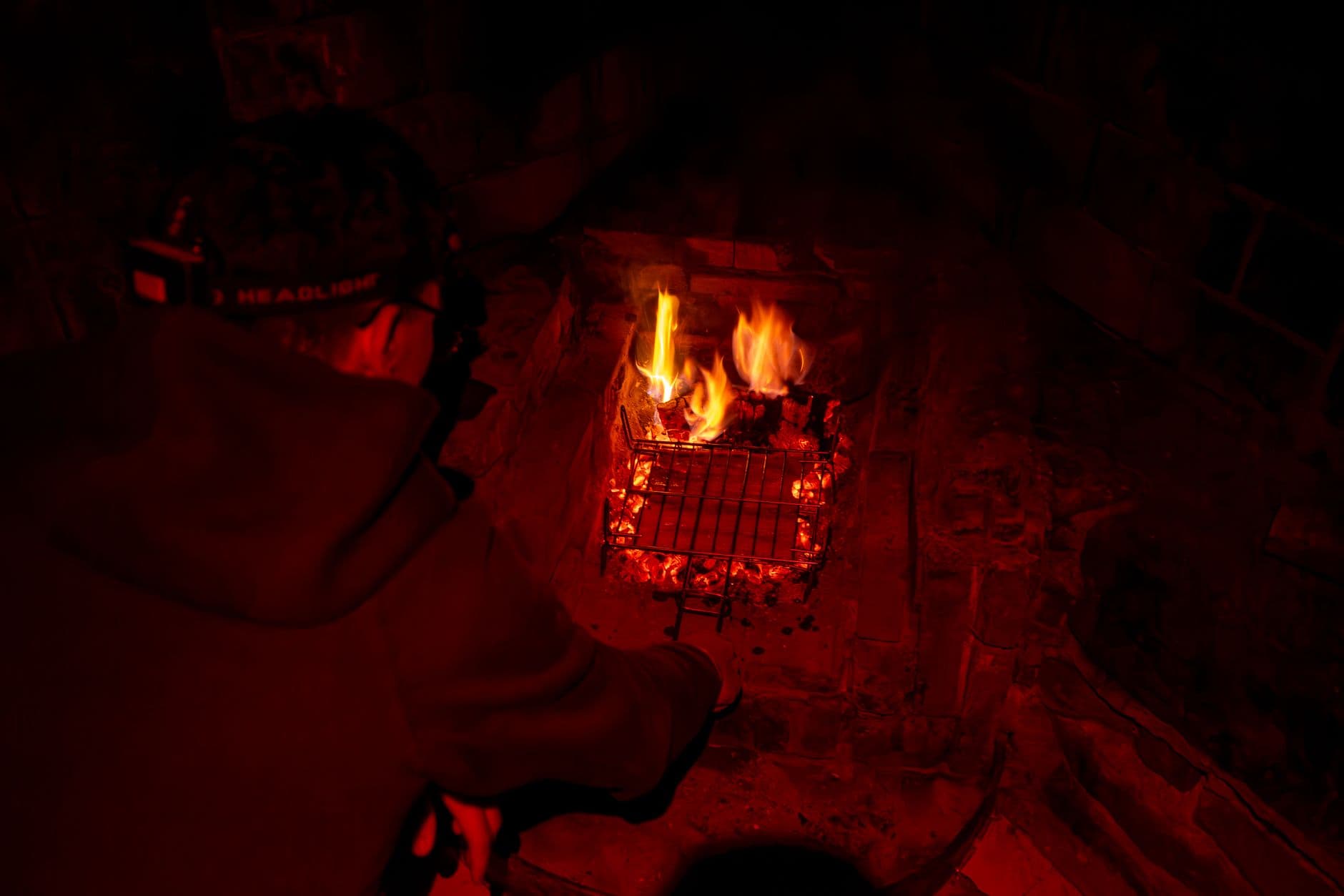 A person cooks over an open fire with a headlamp, creating a cozy campfire ambiance at night.